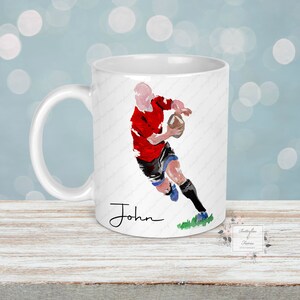 May include: White ceramic mug featuring a watercolor illustration of a rugby player in a red jersey and black shorts, holding a brown ball. The name "John" is written in black script. The mug has a white handle and a green patch of grass.