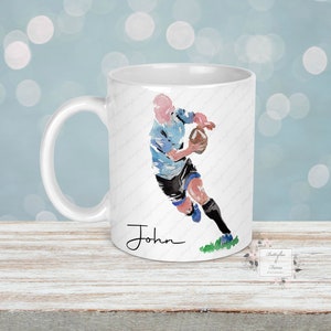 May include: White ceramic mug with a blue and black watercolor illustration of a rugby player running with the ball. The mug has a black handle and the name "John" is printed on the bottom.