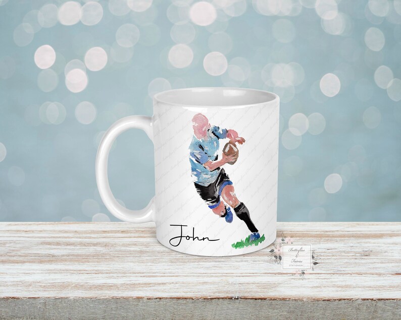 May include: White ceramic mug with a blue and black watercolor illustration of a rugby player running with the ball. The mug has a black handle and the name "John" is printed on the bottom.