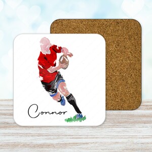 May include: A square coaster with a watercolor illustration of a rugby player in a red jersey and black shorts, holding a brown ball. The name "Connor" is written in cursive. A cork coaster is next to it.
