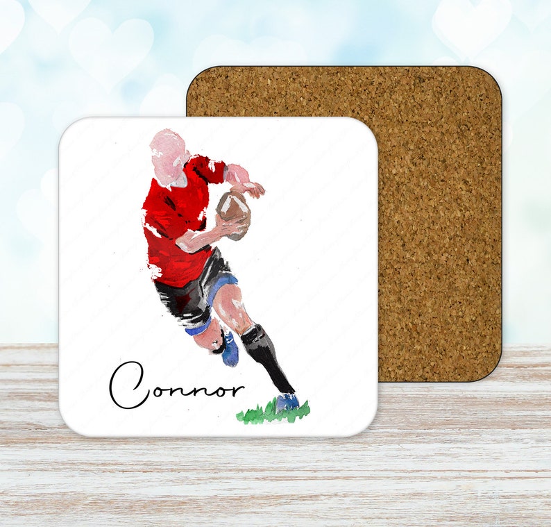 May include: A square coaster with a watercolor illustration of a rugby player in a red jersey and black shorts, holding a brown ball. The name "Connor" is written in cursive. A cork coaster is next to it.