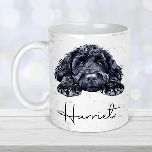 May include: White ceramic mug featuring a black dog illustration with brown eyes and the name "Harriet" in cursive. The dog's paws are visible, resting on the mug's surface. The mug has a white handle and a glossy finish.