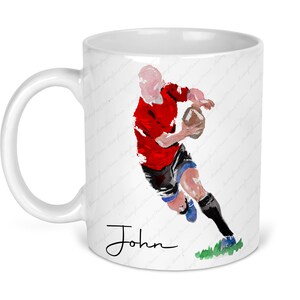 May include: White ceramic mug featuring a watercolor illustration of a rugby player in a red jersey, black shorts, and blue socks, holding a brown ball. The name "John" is written in black script. The mug has a curved handle.
