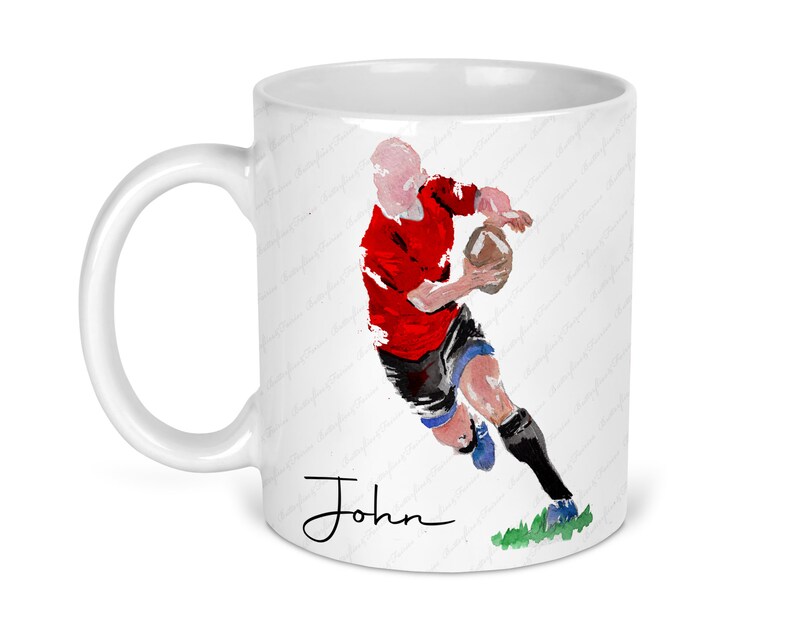 May include: White ceramic mug featuring a watercolor illustration of a rugby player in a red jersey, black shorts, and blue socks, holding a brown ball. The name "John" is written in black script. The mug has a curved handle.