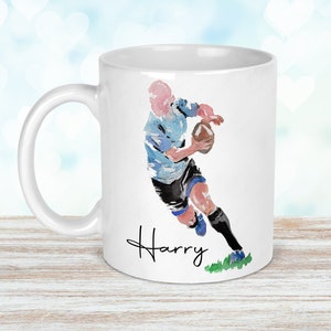May include: White ceramic mug with a watercolor illustration of a rugby player running with the ball. The mug has the name "Harry" written in black cursive at the bottom.