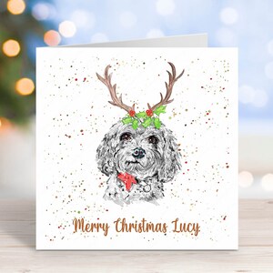 May include: A white Christmas card featuring a watercolour illustration of a dog wearing reindeer antlers and a red bow. The card has a white background with red, green and gold glitter. The text "Merry Christmas Lucy" is printed in brown at the bottom of the card.