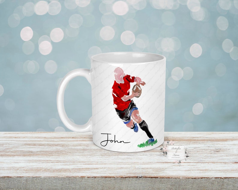May include: White ceramic mug featuring a watercolor illustration of a rugby player in a red jersey and black shorts, holding a brown ball. The name "John" is written in black script. The mug has a white handle and a green patch of grass.