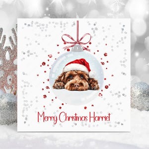 May include: A festive Christmas card with a brown dog in a Santa hat inside a bauble. The card has a white background with silver and red glitter, and the text "Merry Christmas Harriet" in red.