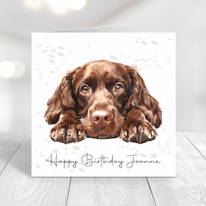 May include: A birthday card featuring a brown dog with its head resting on its paws. The card has a white background with a subtle pattern of paw prints and the text "Happy Birthday Joanne".