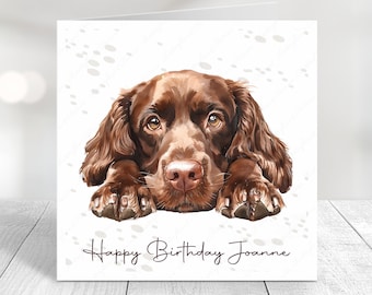 May include: A birthday card featuring a brown dog with its head resting on its paws. The card has a white background with a subtle pattern of paw prints and the text "Happy Birthday Joanne".