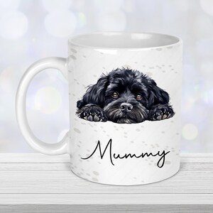 May include: White ceramic mug featuring a black dog illustration with brown eyes and the word "Mummy" in a script font. The mug has a white handle and is decorated with paw prints.