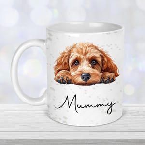 May include: White ceramic mug with a brown and tan dog illustration and the word "Mummy" in script. The dog rests its paws on the mug's edge. The mug has a white handle and is decorated with paw prints. A charming gift.