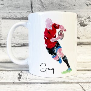 May include: White ceramic mug featuring a watercolor illustration of a rugby player in a red jersey, black shorts, and blue socks. The mug has a curved handle and the name "Guy" is written in cursive.