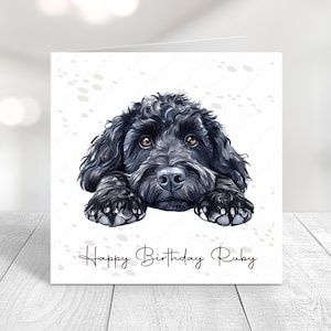 May include: A square birthday card with a detailed illustration of a black dog with brown eyes. The dog's paws are resting on the bottom edge of the card. The words "Happy Birthday Ruby" are written in cursive below the dog.
