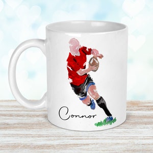 May include: White ceramic mug featuring a watercolor illustration of a rugby player in a red jersey, black shorts, and blue socks, holding a football. The name "Connor" is written in cursive below the image.