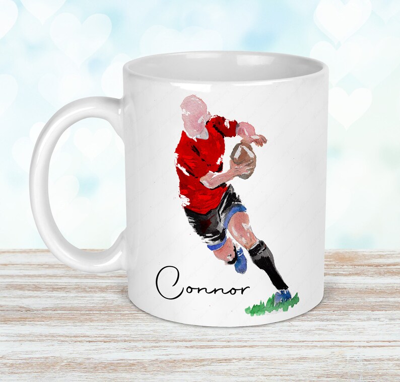 May include: White ceramic mug featuring a watercolor illustration of a rugby player in a red jersey, black shorts, and blue socks, holding a football. The name "Connor" is written in cursive below the image.