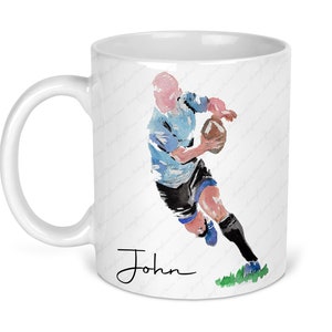 May include: White ceramic mug with a watercolor illustration of a rugby player running with the ball. The player is wearing a blue and white jersey and black shorts. The mug has the name "John" written in black cursive at the bottom.