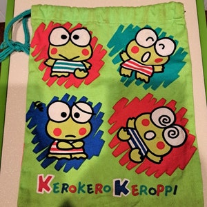 May include: Green drawstring bag with four cartoon frogs wearing striped swimsuits. The frogs are surrounded by colorful scribbles. The bag has the text "Kero Kero Keroppi" printed on the bottom.