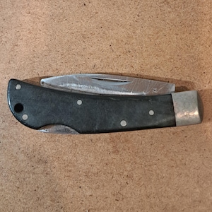 May include: A black folding pocket knife with a silver blade and accents. The knife has a dark handle with three silver rivets and a silver end cap. The blade is partially open, revealing its sharp edge. The knife is on a brown surface.