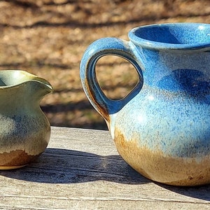 May include: Two ceramic pitchers, one blue and one green, with a brown glaze. The pitchers are both round and have a handle on one side.
