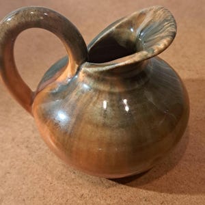 May include: A small, round ceramic pitcher featuring a glossy glaze with blended tones of earthy brown, deep charcoal, and warm orange. The vessel has a curved handle on one side and a wide, flared pouring spout at the top of the rounded body.