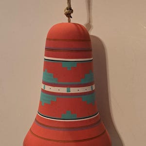 May include: A hand-painted, red ceramic bell with turquoise and white geometric patterns. The bell is suspended by a natural rope and has a black clapper. The bell is a decorative item.