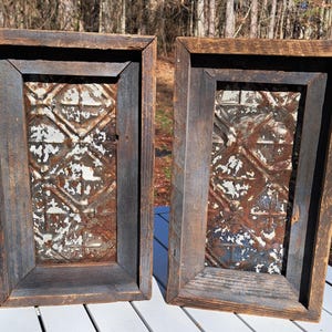 May include: Two rectangular wooden framed wall decor pieces. The frames are made of dark, weathered wood. The center panels feature a distressed, patterned metal design with white and rust-colored accents. The pieces are displayed outdoors.