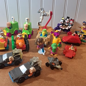 May include: A collection of colorful toy figures and vehicles featuring cartoon characters. Includes Bugs Bunny, the Warner Bros., Barney, Dora the Explorer, and characters from Back to the Future. The toys are made of plastic and are in various sizes.