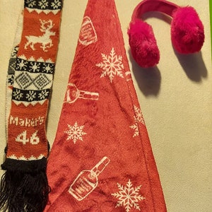 May include: A red Santa hat with a white pom-pom on top. The hat has a white snowflake and bottle pattern. A black and orange knitted scarf with the text "Maker's 46" and a pair of pink fuzzy earmuffs are also included.
