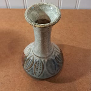 May include: A ceramic vase with a flared rim and a narrow neck, featuring a speckled off-white glaze on the upper portion and a base decorated with a repeating pattern of brown and grey leaf-like shapes against a light-colored background.