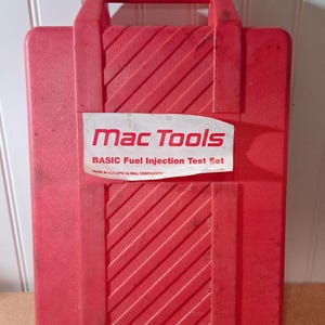 May include: A red plastic carrying case with a handle, featuring a white label that reads Mac Tools BASIC Fuel Injection Test Set MADE IN USA WITH GLOBAL COMPONENTS. The case has a textured surface with diagonal raised lines on the front panel.