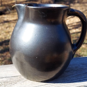 May include: A rounded ceramic pitcher with a dark, matte black finish and a subtle metallic sheen. The vessel features a curved handle on the right side, a wide body, and a small, defined pouring spout at the top rim, showcasing a smooth texture.