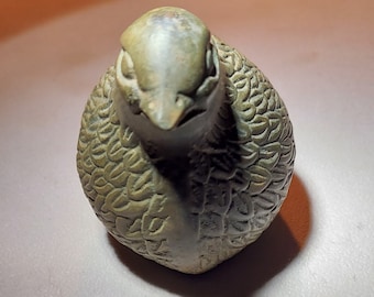 May include: A small, green ceramic bird figurine with detailed feather markings. The bird is facing down with its head tilted slightly to the side.