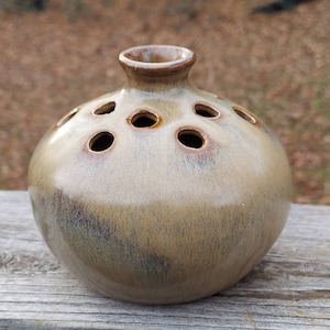 May include: A round, light brown ceramic vase with a textured glaze and multiple holes around the top for flower stems.