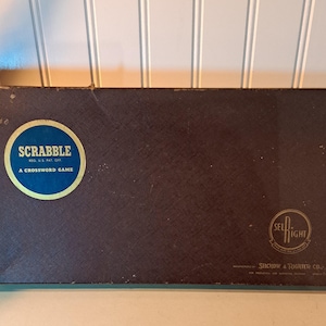 May include: A brown cardboard box with the words "SCRABBLE" and "A CROSSWORD GAME" printed on the front. The box also has the Selchow & Righter Co. logo.