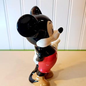 May include: A black and white ceramic figurine of Mickey Mouse wearing red shorts and brown shoes. The figurine is standing with its right arm raised.