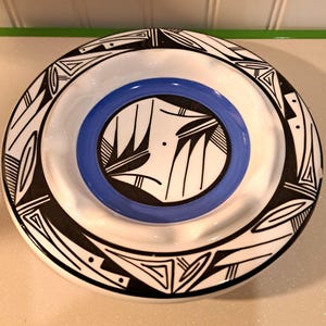 May include: A round, white ceramic ashtray with a blue center and black geometric designs. The ashtray features a central blue ring and a decorative border with black and white patterns. The design is reminiscent of Native American pottery.