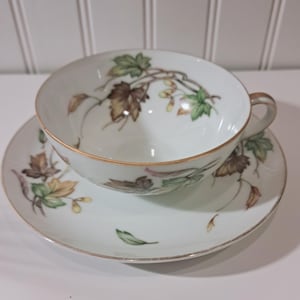 May include: White teacup and saucer set with a gold rim and a painted leaf design in shades of green, brown, and yellow. The teacup has a handle and sits on a matching saucer. A classic tea set.