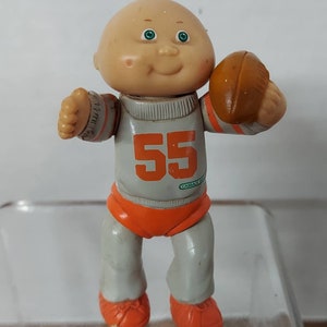 May include: A Cabbage Patch Kid doll wearing a gray and orange football uniform with the number 55 on the front. The doll is holding a brown football and has a bald head with green eyes.