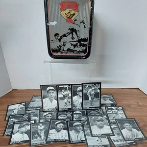 May include: A collection of black and white baseball trading cards featuring images of famous baseball players. The cards are arranged in a pile on a wooden surface. A metal tin with a baseball scene is in the background.