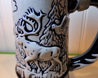 May include: A white ceramic beer stein with a blue and brown rim. The stein features a detailed illustration of an Alaskan moose in a forest setting. The text "Alaskan Moose" is visible on the bottom of the stein.