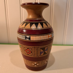 May include: A small, brown ceramic vase with geometric patterns in shades of orange, red, green, blue, and white. The vase has a narrow neck and a rounded body.
