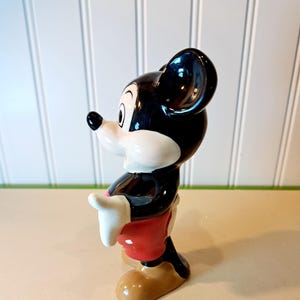 May include: A ceramic figurine of Mickey Mouse. He is standing with his right hand raised and wearing red shorts with white polka dots. His left hand is on his hip. He has black shoes and a black and white face.