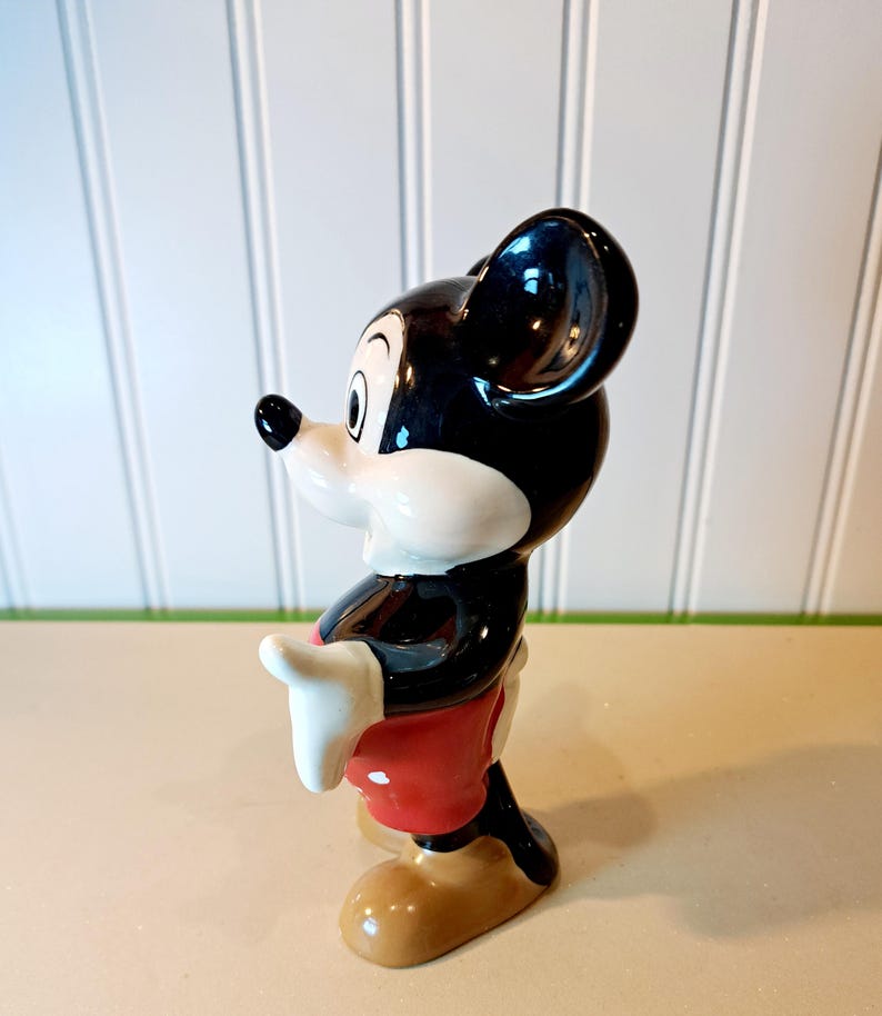 May include: A ceramic figurine of Mickey Mouse. He is standing with his right hand raised and wearing red shorts with white polka dots. His left hand is on his hip. He has black shoes and a black and white face.