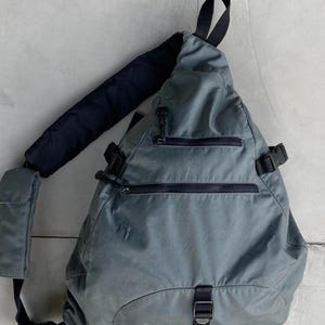 May include: A gray sling backpack with black zippers and straps. The bag has a padded shoulder strap and a small side pocket. The design is minimalist and functional, suitable for everyday use.