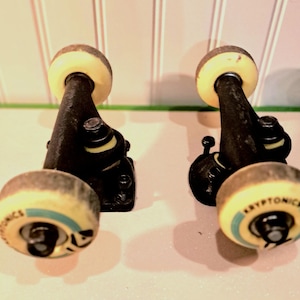 May include: Two black skateboard trucks with yellow and blue wheels. The wheels have the word "Kryptonics" printed on them. The trucks are set against a light background.