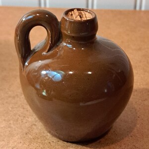 May include: A small, round, brown glazed ceramic jug featuring a single side handle and a narrow neck opening. The top of the jug is sealed with a light brown natural cork stopper. The surface of the ceramic has a smooth, glossy, reflective finish.