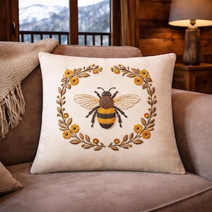 Puede incluir: Cojín cuadrado crema con un diseño de abeja bordada. La abeja es marrón y amarilla, rodeada por una corona floral en tonos amarillos y marrones. El cojín está sobre un sofá marrón.