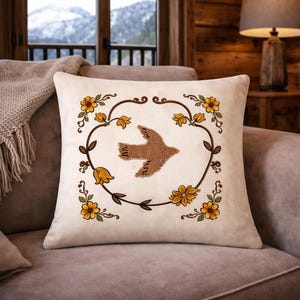 Puede incluir: Cojín cuadrado crema con un pájaro bordado marrón en el centro. El pájaro está rodeado por una corona floral marrón con flores amarillas y naranjas. El cojín está sobre un sofá marrón.