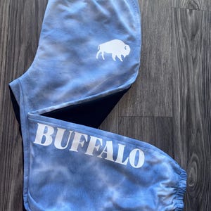 Pode incluir: Calças de moletom azuis claras com efeito tie-dye com um gráfico de búfalo branco na perna superior esquerda e a palavra "BUFFALO" impressa em branco na perna inferior. As calças de moletom têm cós e punhos elásticos.